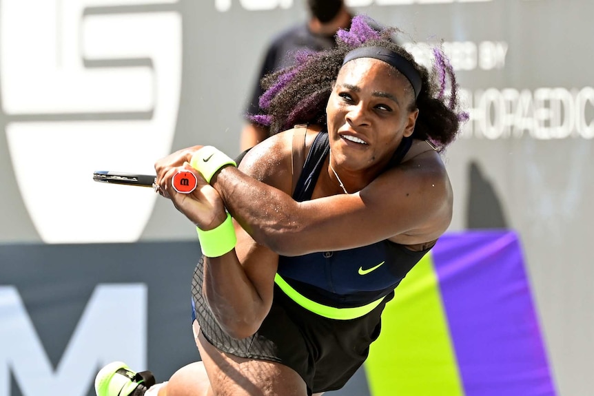 Serena Williams finishes playing a shot in a black and green outfit wearing a headband and purple streaks in her hair
