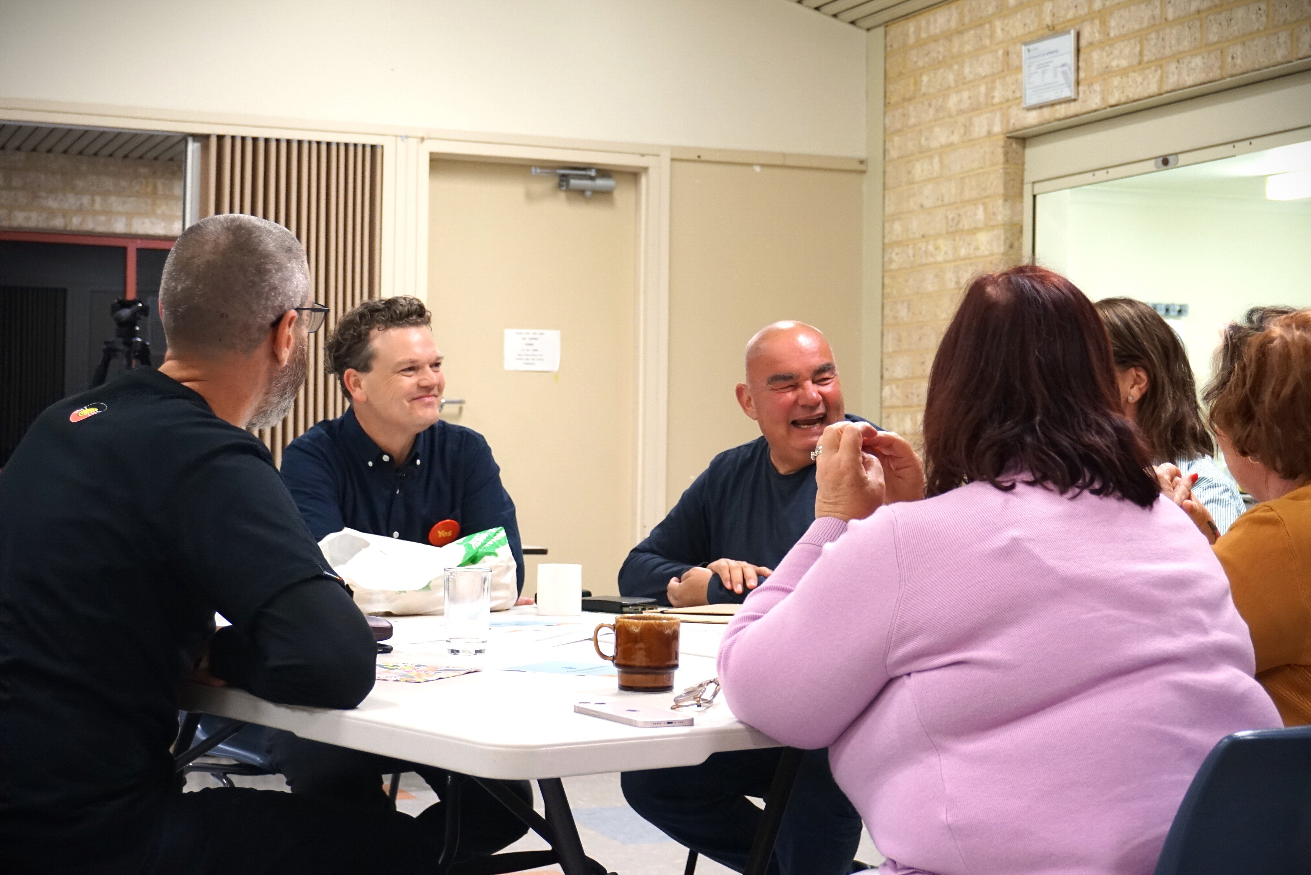 A group of six people sit around a table holding a discussion, with coffee cups and phones among the items on the table.