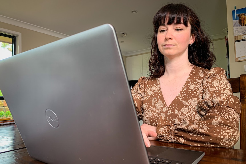 A woman in a brown dress sits at a desk working on a laptop