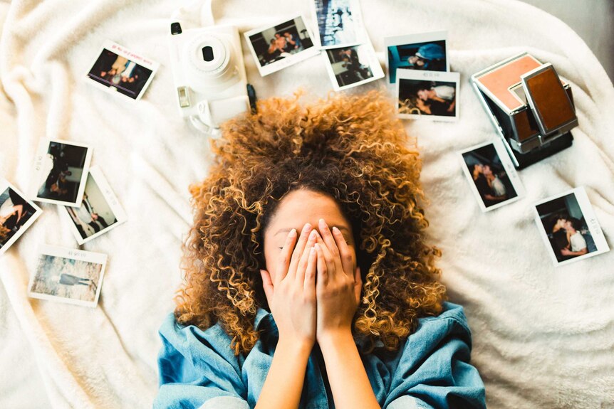 A woman with hand covering her face lying on a bed surrounded by photos