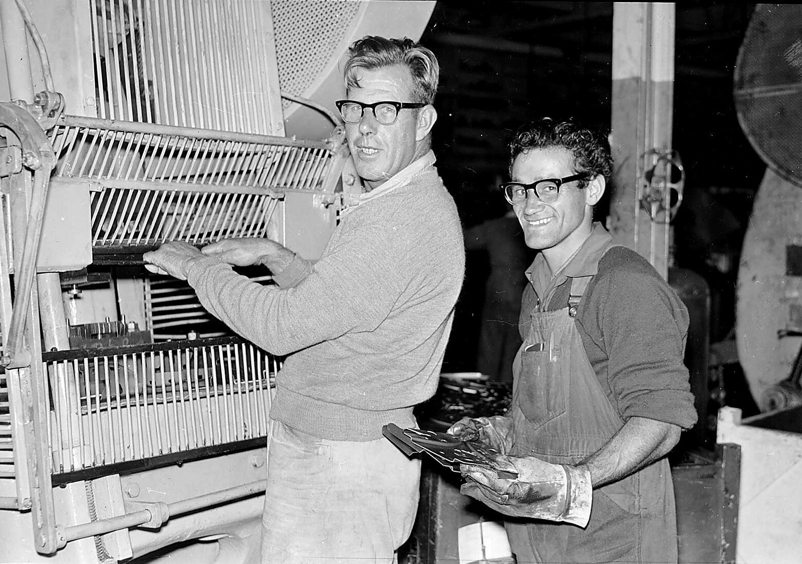 A black and white photo of two men, in overalls and jumpers working on a machine