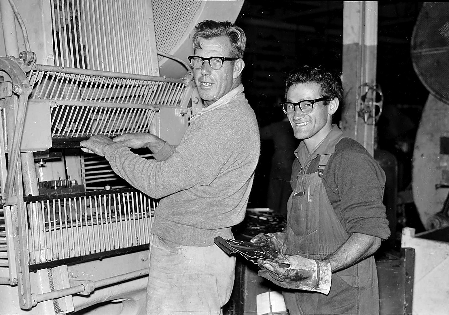 A black and white photo of two men, in overalls and jumpers working on a machine