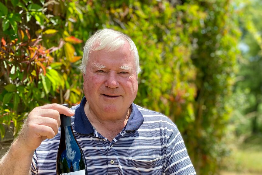 Man with white hair holds wine bottle in front of vibrant green and red vines