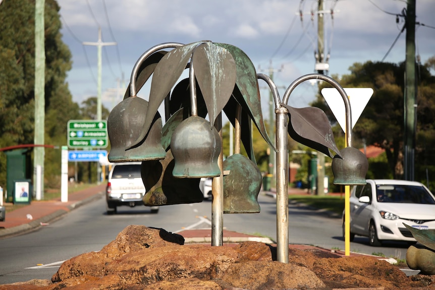 An image of a metal gumnut statue at a roundabout during an overcast day. Cars in the background and signs for suburbs.
