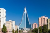 Colour photograph of a road leading towards Ryugyong Hotel towering over the Pyongyang cityscape in front of a clear blue sky.