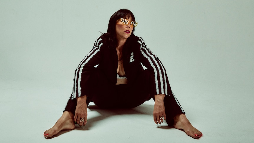 Katy Steele sits on ground barefoot wearing dark tracksuit and gold glasses that say 'BIG STAR'