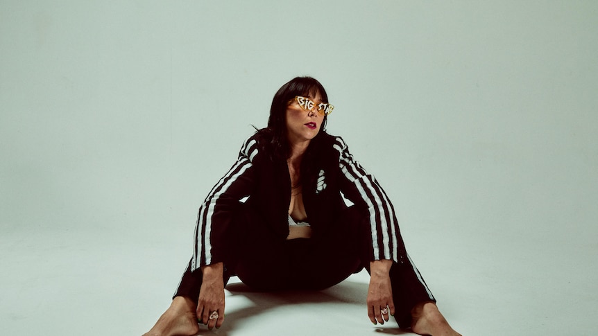 Katy Steele sits on the floor in a black tracksuit with sunglasses that say 'Big Star'