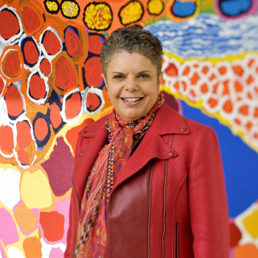 Deborah Cheetham, standing in front of a brightly coloured painting, smiles at the camera.