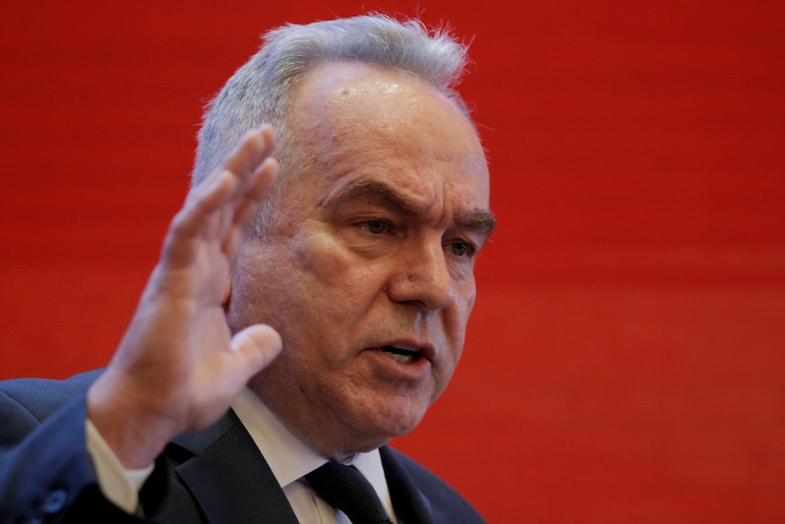 A man in a suit holds up his hand in front of a red backdrop