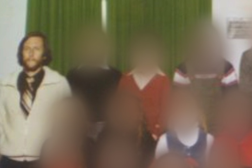 A class photo with a male teacher with a beard standing far left. All other faces are blurred.