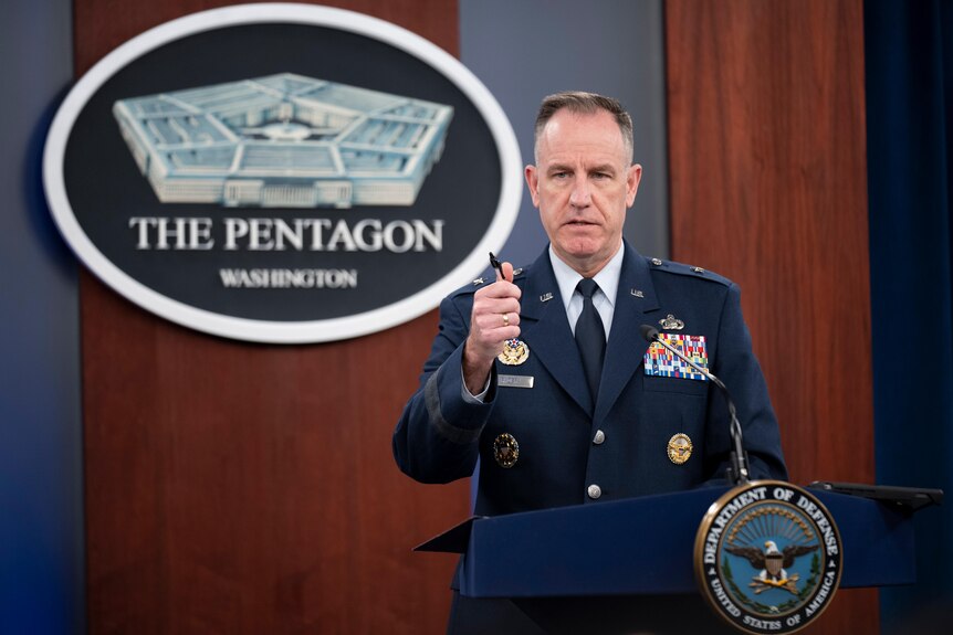 An Air Force general in dress uniform points as he speaks from behind a lecturn in a formal meeting room with a Pentagon sign.