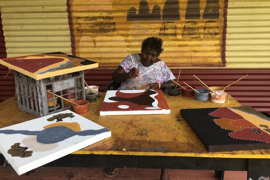 A woman sits at a table painting while three unfinished canvas sit around her