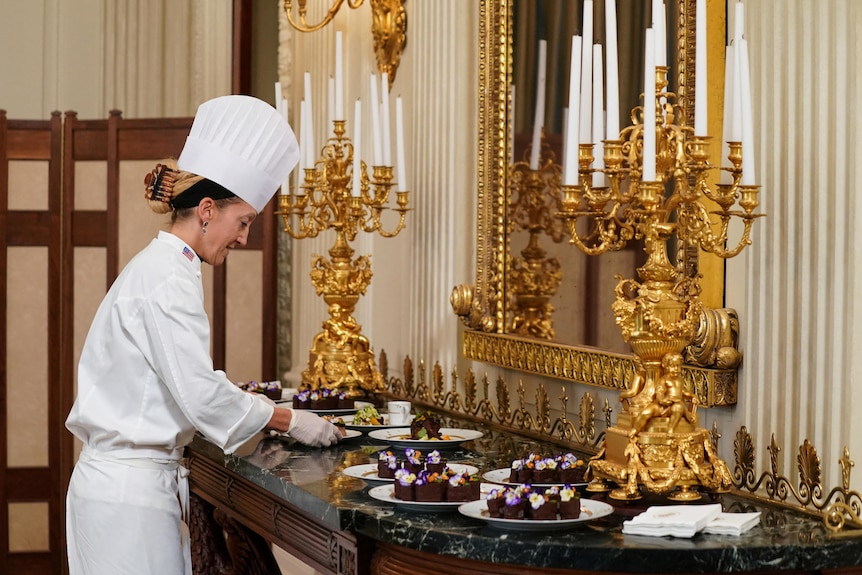 A chef wearing a white hat and gloves places a plate on an ornate bar