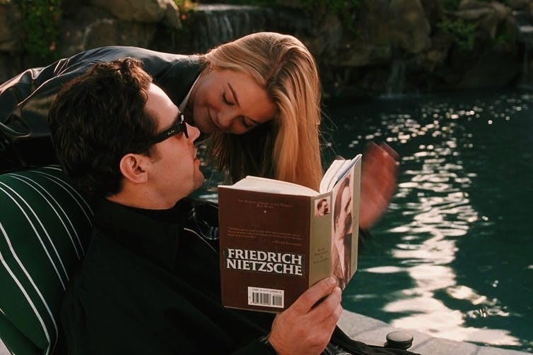 A preppy girl leans over a man in Raybans reading Nietzsche