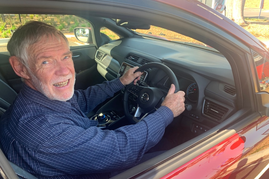 A man with grey hair and a grey beard sitting in a red car with his hands on the steering wheel.