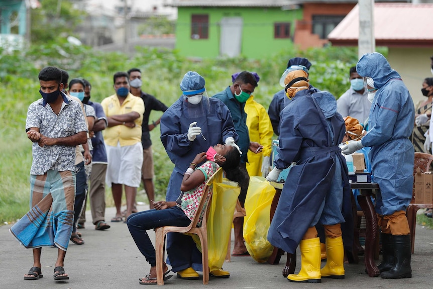 A group of men wait in a line with masks on as a woman sits in a chair as a man in scrubs moves to swab her nose