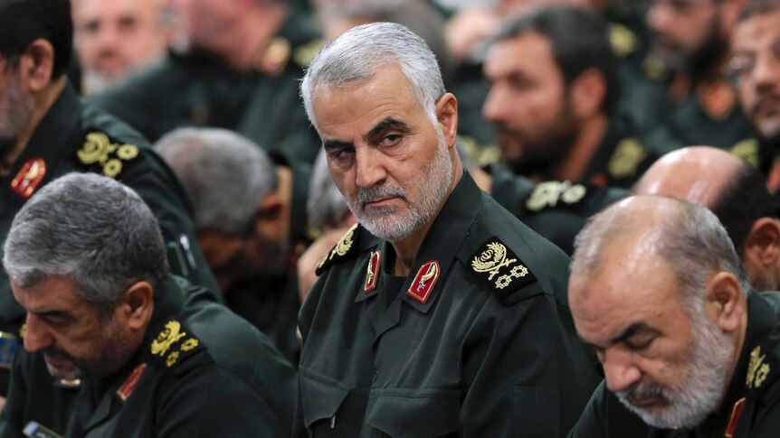 General Qassem Soleimani in military uniform with grey hair and dark eyebrows glances from crowd of military men.