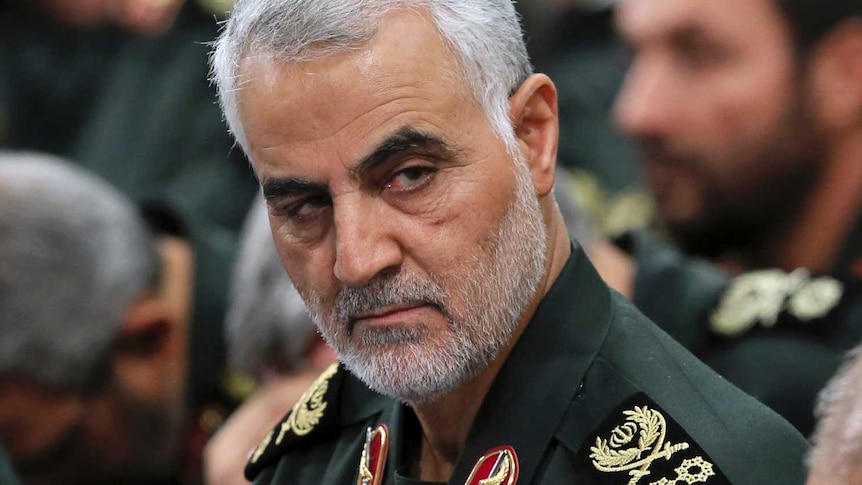 General Qassem Soleimani in military uniform with grey hair and dark eyebrows glances from crowd of military men.