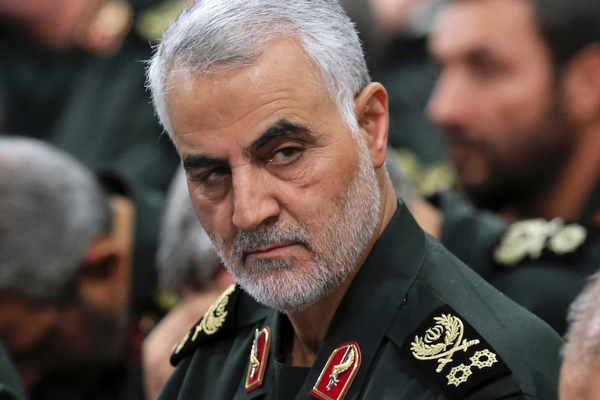 General Qassem Soleimani in military uniform with grey hair and dark eyebrows glances from crowd of military men.