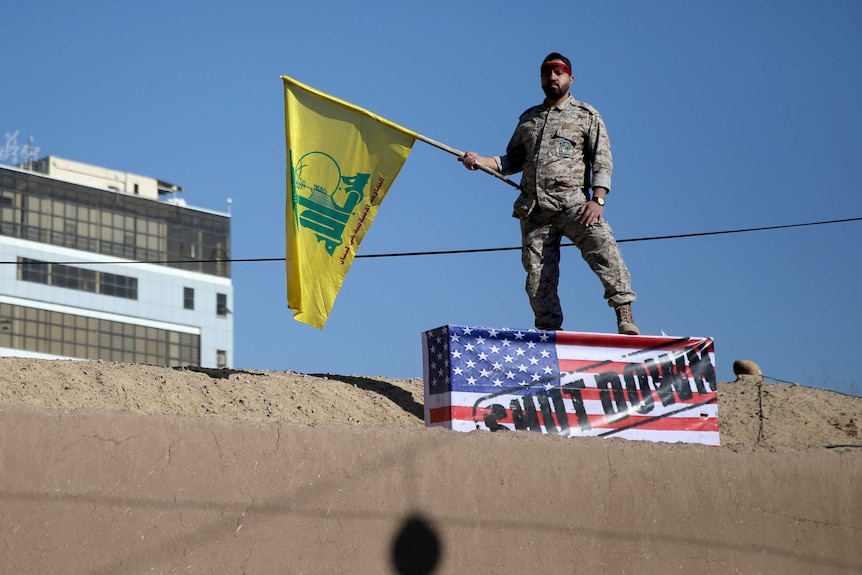 An Iranian soldier stands on a box with a US flag while he holds the yellow Hezbollah flag on top of a stone wall on a clear day