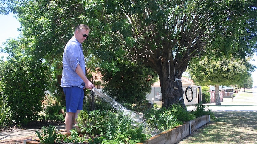 Man stands hosing a raised garden bed on a footpath.