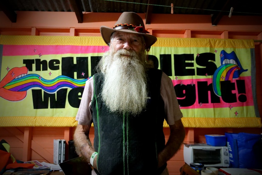 A man with a long white beard and broad brim hat in front of a colourful poster