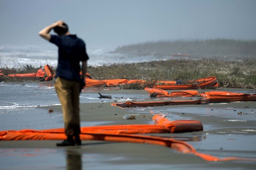 A television report stands with his hands on his head beside oil booms on a beach stained with oil.