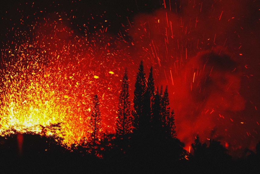 Red hot lava flies across into the air near trees during the night.