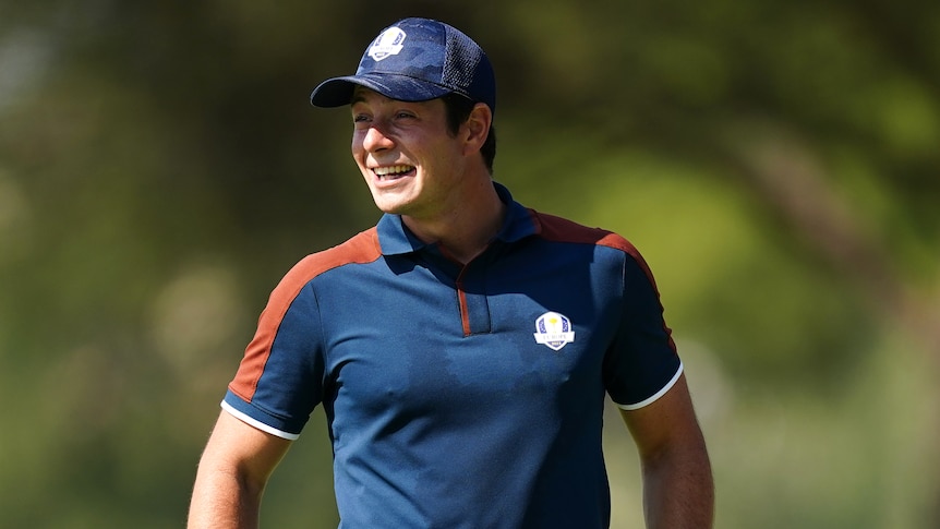 A clean-shaven man wearing a blue polo shirt and cat smiles. 
