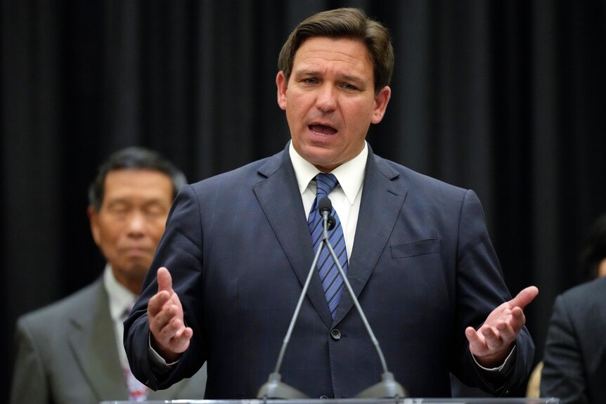 A middle-aged white man with brown hair wearing a suit gestures with both hands as he speaks at a podium.