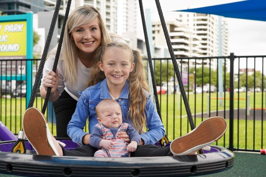 Stephanie Hewins with her young daughter Lilly, holding a young baby on a rope swing in a park in the middle of a city.