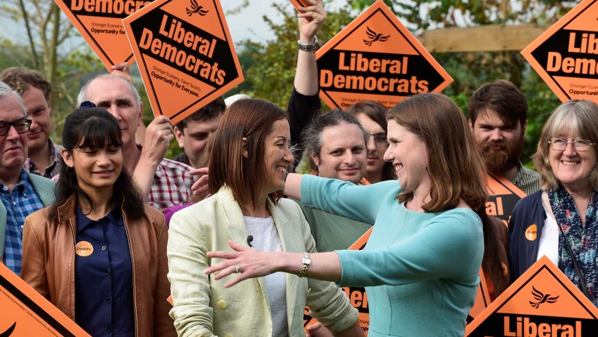 Two women hug in front of political party supporters holding signs.