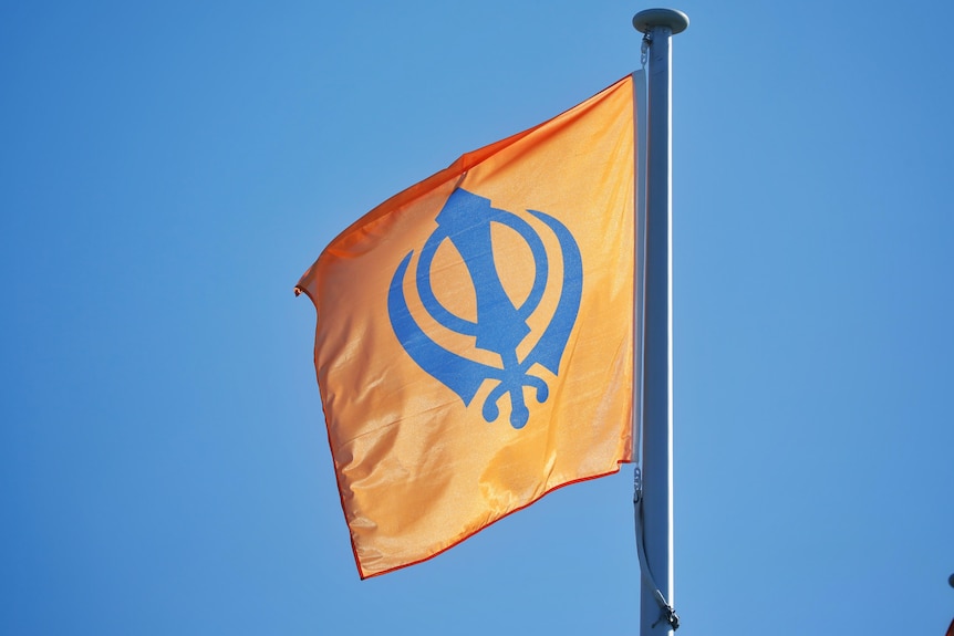 A close shot of an orange and blue Sikh flag flying on a flagpole in front of a bright blue sky.
