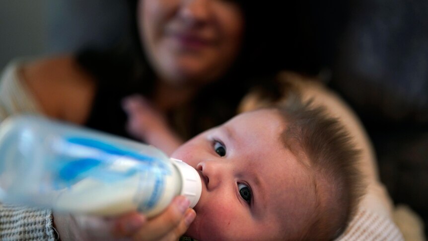 a close up of a baby drinking from a bottle looking at the camera as the child's mother holds him