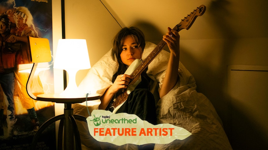 Sophie May sits in a beanbag in a room lit by a small white lamp, holding her guitar.