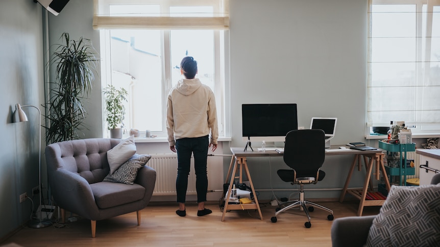 A man stands next to a computer desk in his living room while looking out the window