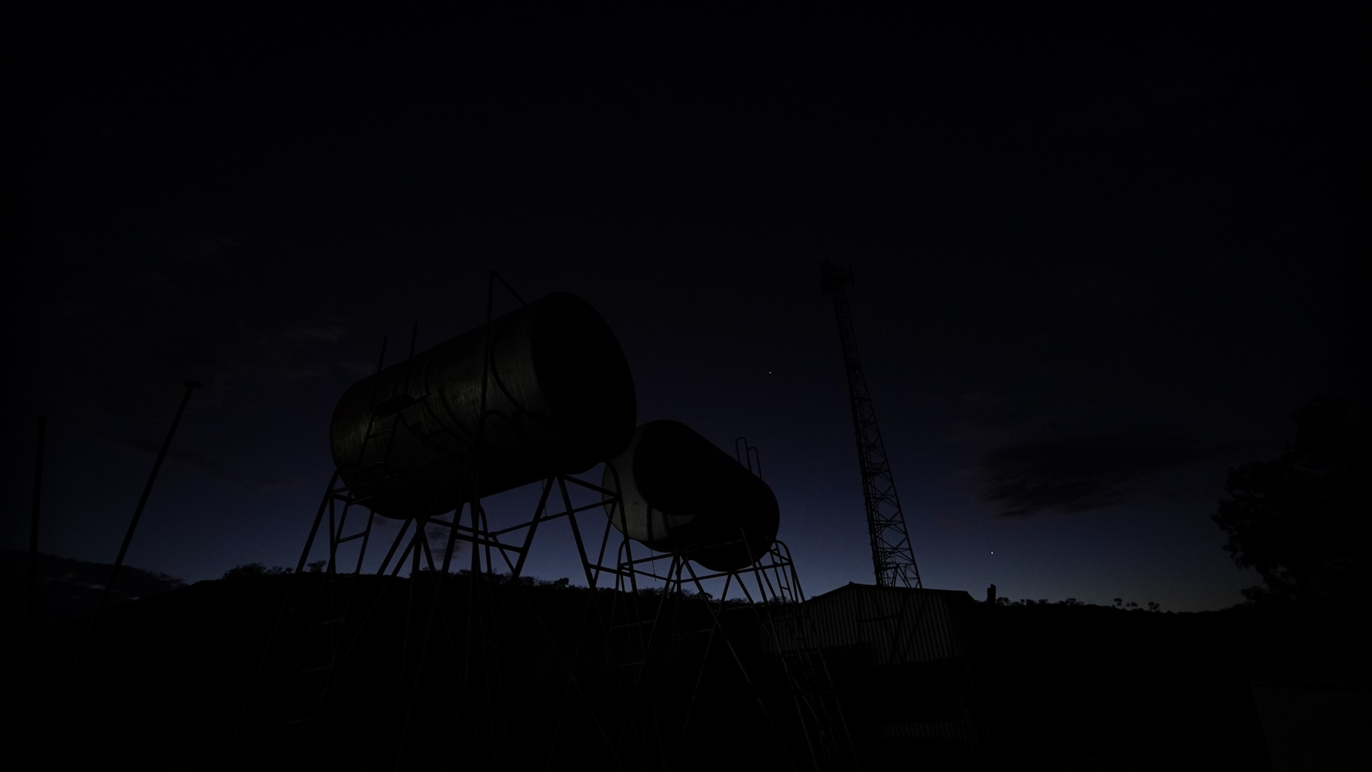 Metal structures at a remote roadhouse and rocky landscapes are silhouetted against the darkening night sky behind.