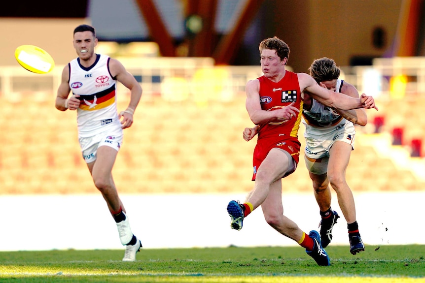 A red-headed AFL footballer kicks the ball flor a long goal for his team as defenders watch on.