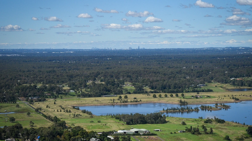 A view of mainly bushland with green paddocks and large dams in the foreground and skyscrapers in the very far distance.