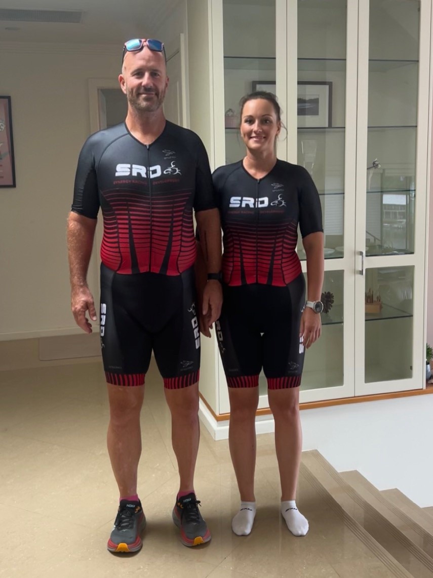 A man and woman stand next to each other inside a house, wearing lycra cycle shorts and shirts.
