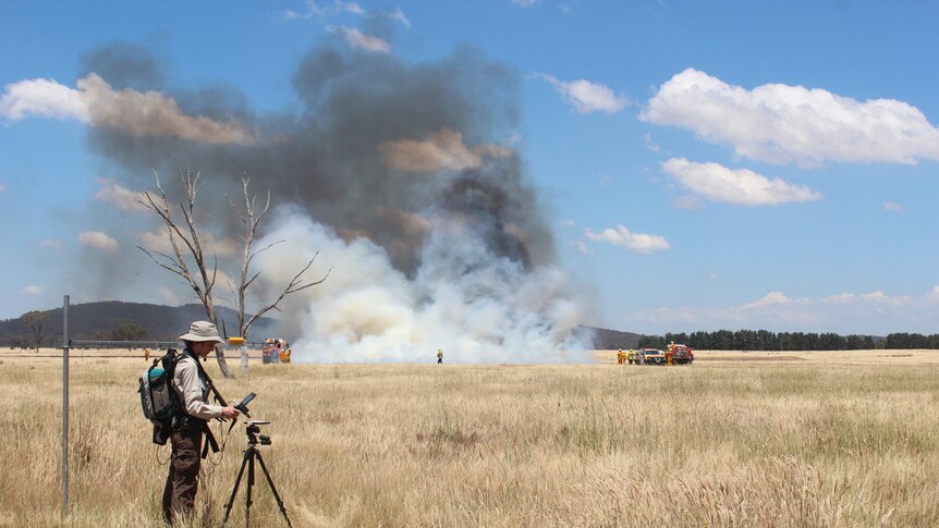 Dr Marta Yebra stands in a field and looking at data, while a controlled fire experiment is underway in the background.