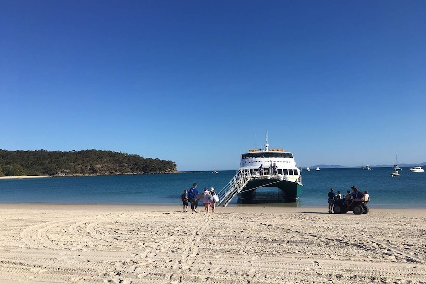 A ferry boat docked on the beach with white sand and blue water as people walk on board.