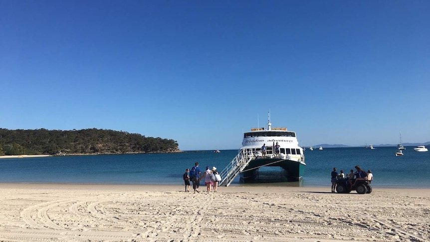 A ferry boat docked on the beach with white sand and blue water as people walk on board.