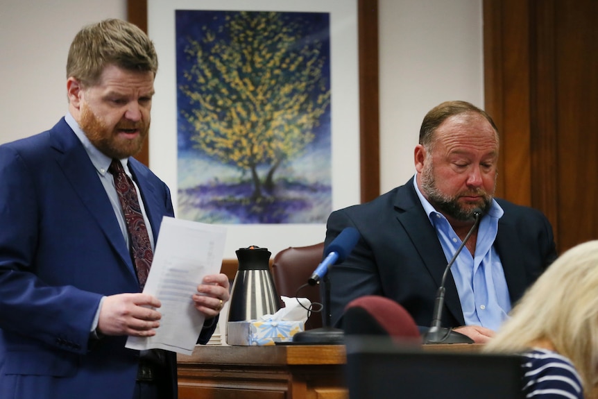 A white man in a courtroom wearing a suit and holding pieces of paper stands next to Alex Jones, who is in the witness box