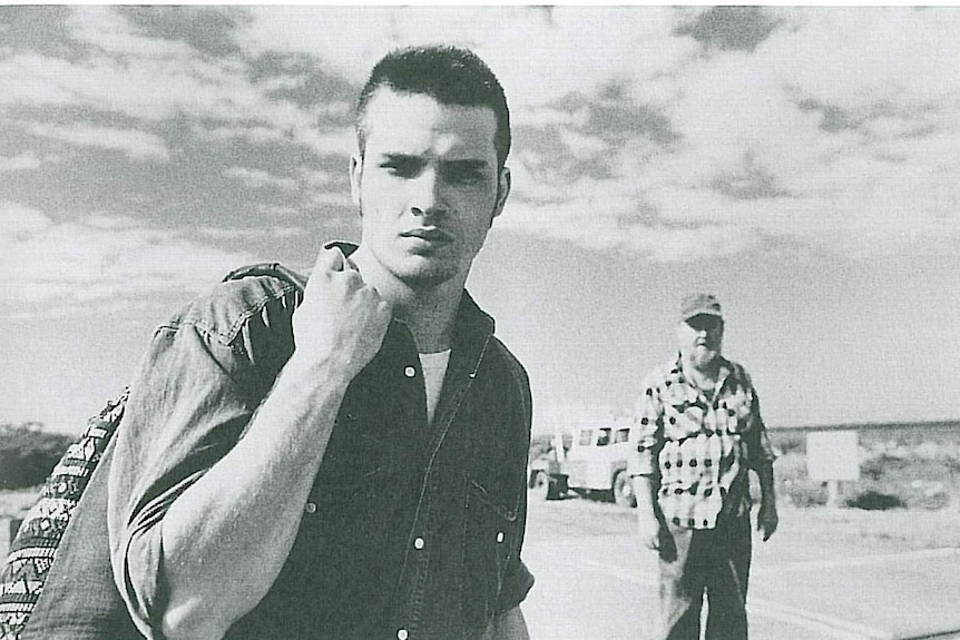Black and white photo of young man standing on a country road, older man with beard in background.