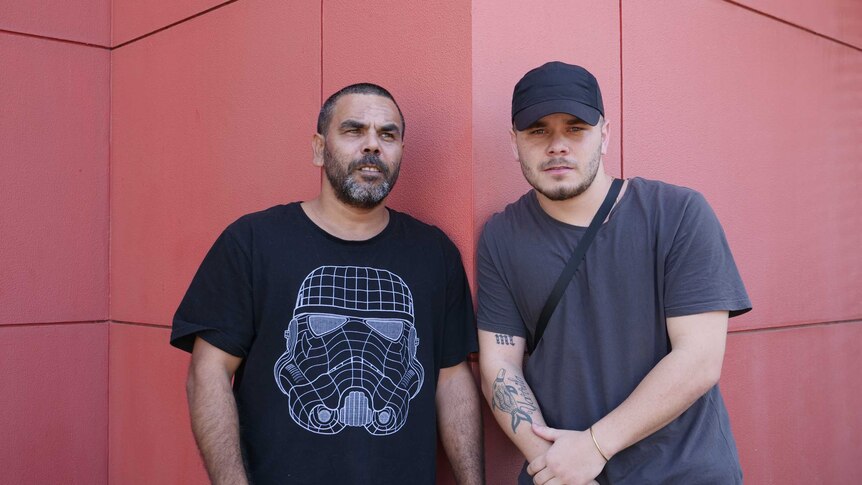 Two Indigenous men standing next to each other with a red wall in background.
