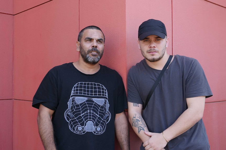 Two Indigenous men standing next to each other with a red wall in background.