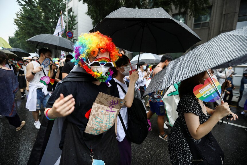 A group of people in a parade one in focus wearing a rainbow wig and lei