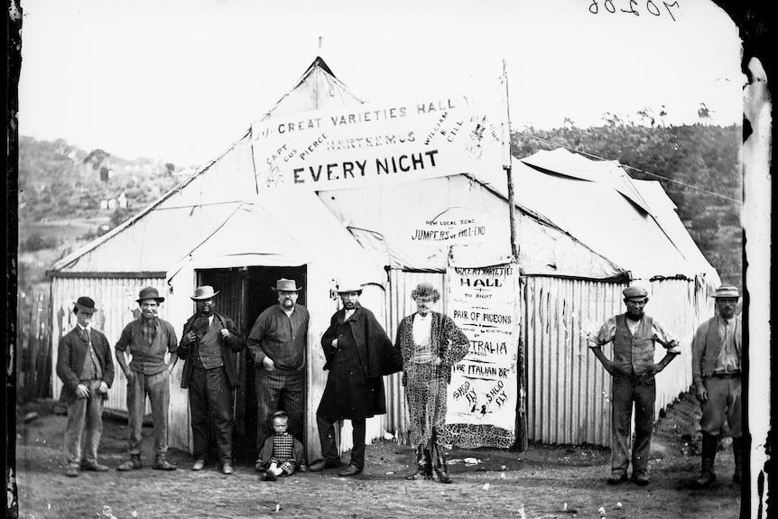 Historical black and white photo of men standing outside the Great Varieties Hall tent theatre in about 1870