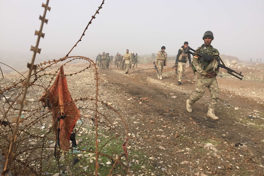 Iraqi Army soldiers walk along a ridge, barbed wire sits in the fore of the image